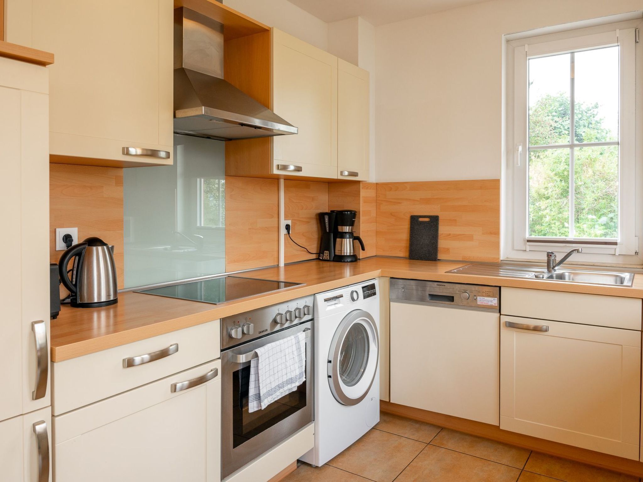 A modern kitchen with wooden cabinets, a stainless steel range hood, a built-in dishwasher, a washing machine, and a window providing natural light.