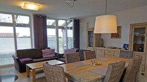 Ein modernes Wohnzimmer mit einer Couch, einem Esstisch und Stühlen. Ein großes Fenster mit Vorhängen, eine Lampe und eine Vitrine mit Gläsern sind ebenfalls vorhanden.