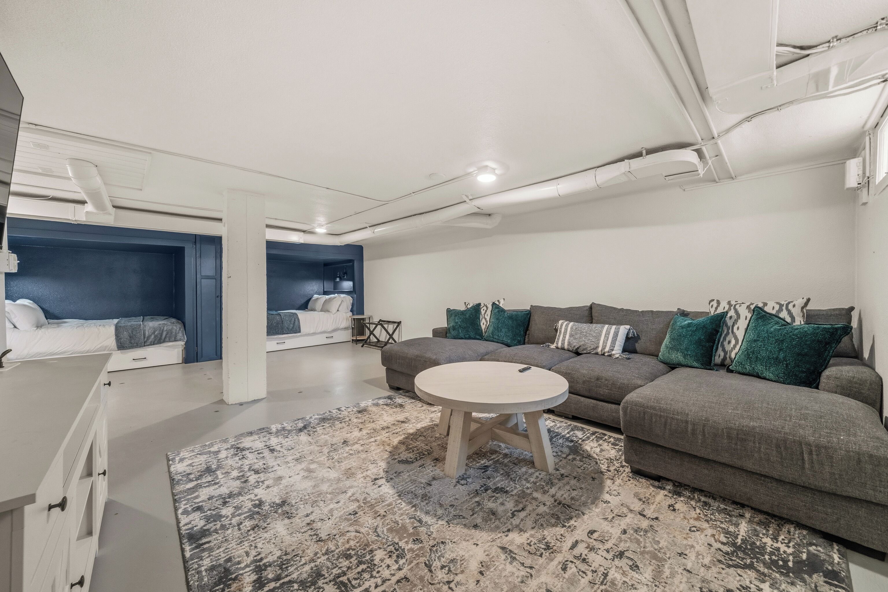 A modern living room with a gray sofa, a round coffee table, and a patterned rug. There is a white cabinet with drawers and a small table with a lamp. The room has a white ceiling with visible pipes and a window on the right side.