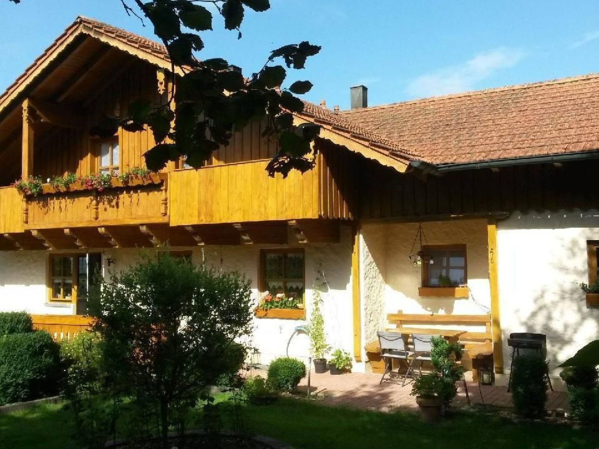 A two-story wooden house with a balcony, a patio with a table and chairs, and a garden area.