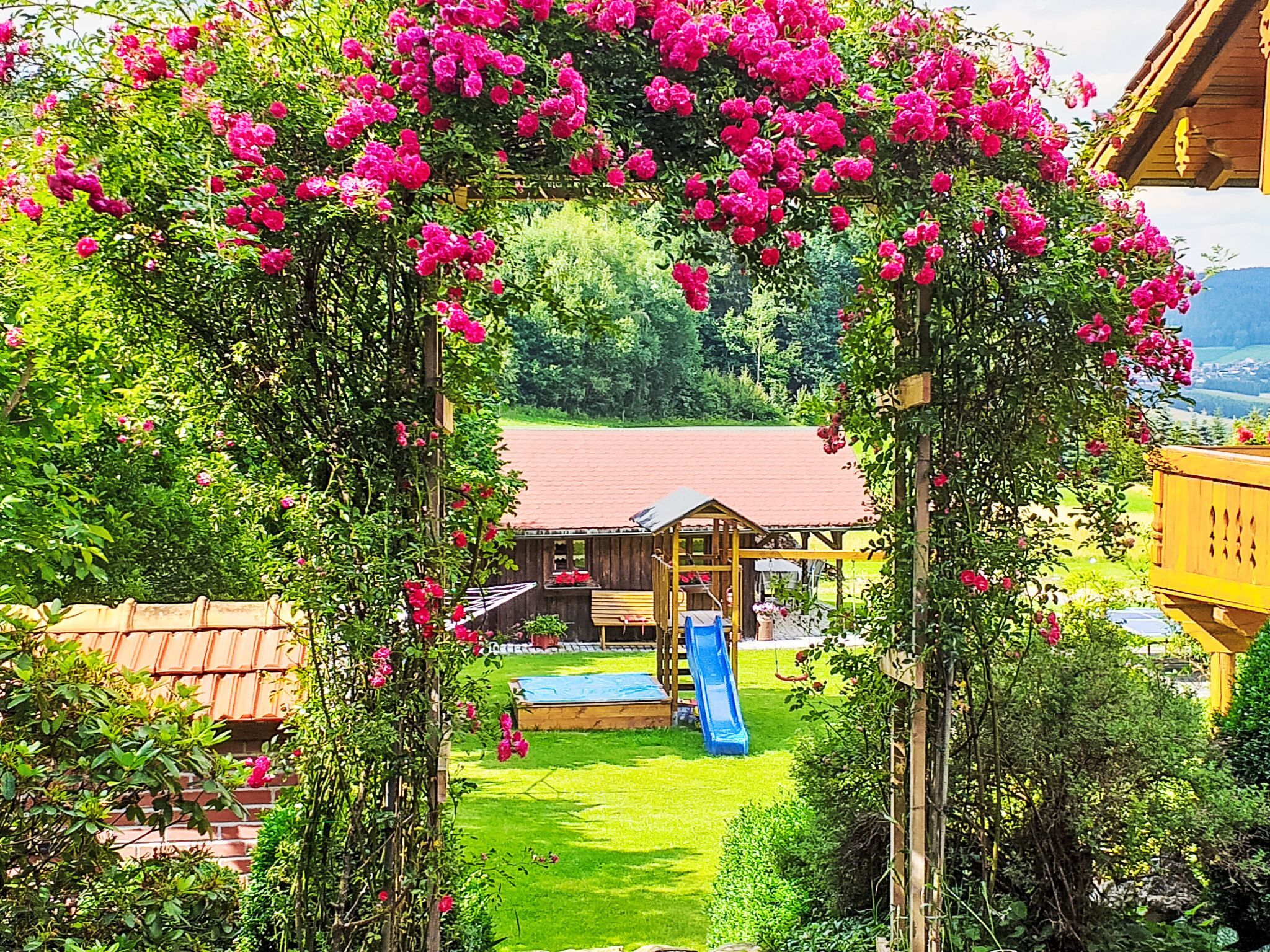 Una casa de fusta amb teulada vermella, tobogan i jardí amb flors roses.