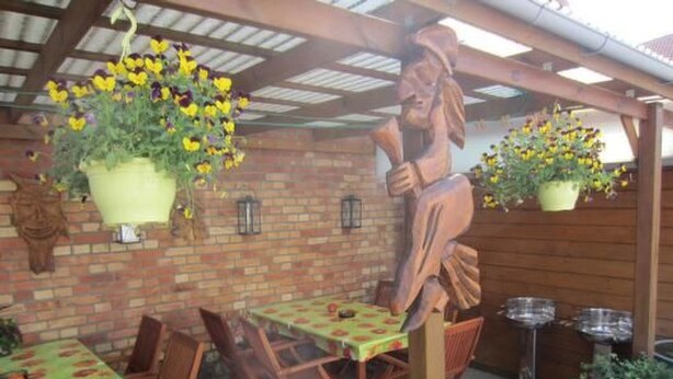 Un patio esterno con parete in mattoni, vasi fioriti pensili, una statua di legno e un tavolo con sedie.