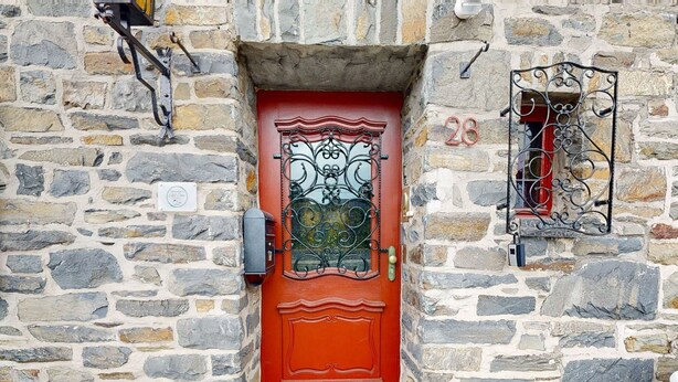 Eine rote Tür mit dekorativem Glasfenster und einem Metallgitter, eingebettet in ein Steinhaus mit einer Steinmauer und einer Hausnummer.