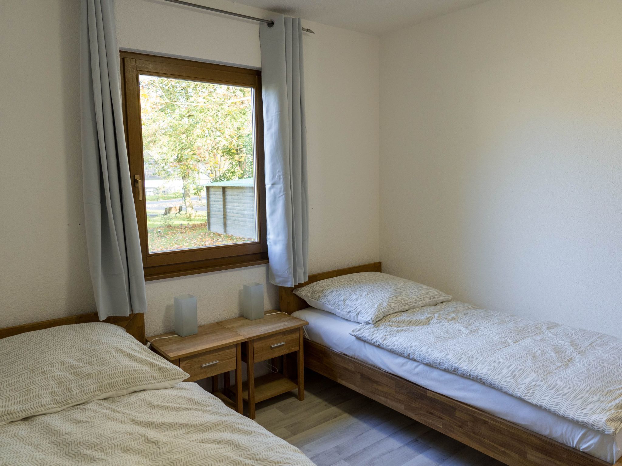 Une petite pièce propre, avec deux lits, un bureau en bois et une fenêtre avec des rideaux.