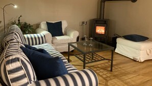 Een gezellige woonkamer met een bank, fauteuil en poef, een glazen salontafel en een houtkachel.
