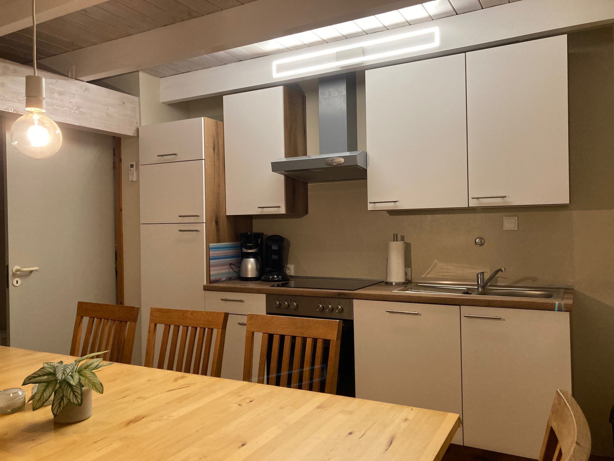 Een compacte keuken met witte kastjes, een roestvrijstalen spoelbak en een houten eettafel met stoelen.