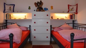 Ein Zimmer mit zwei Betten, beide mit roter Bettdecke und karierten Kissenbezügen. In der Mitte befinden sich weiße Kommoden mit roten Knöpfen. Über den Betten sind Regale mit Büchern und kleinen Dekoartikeln zu sehen.