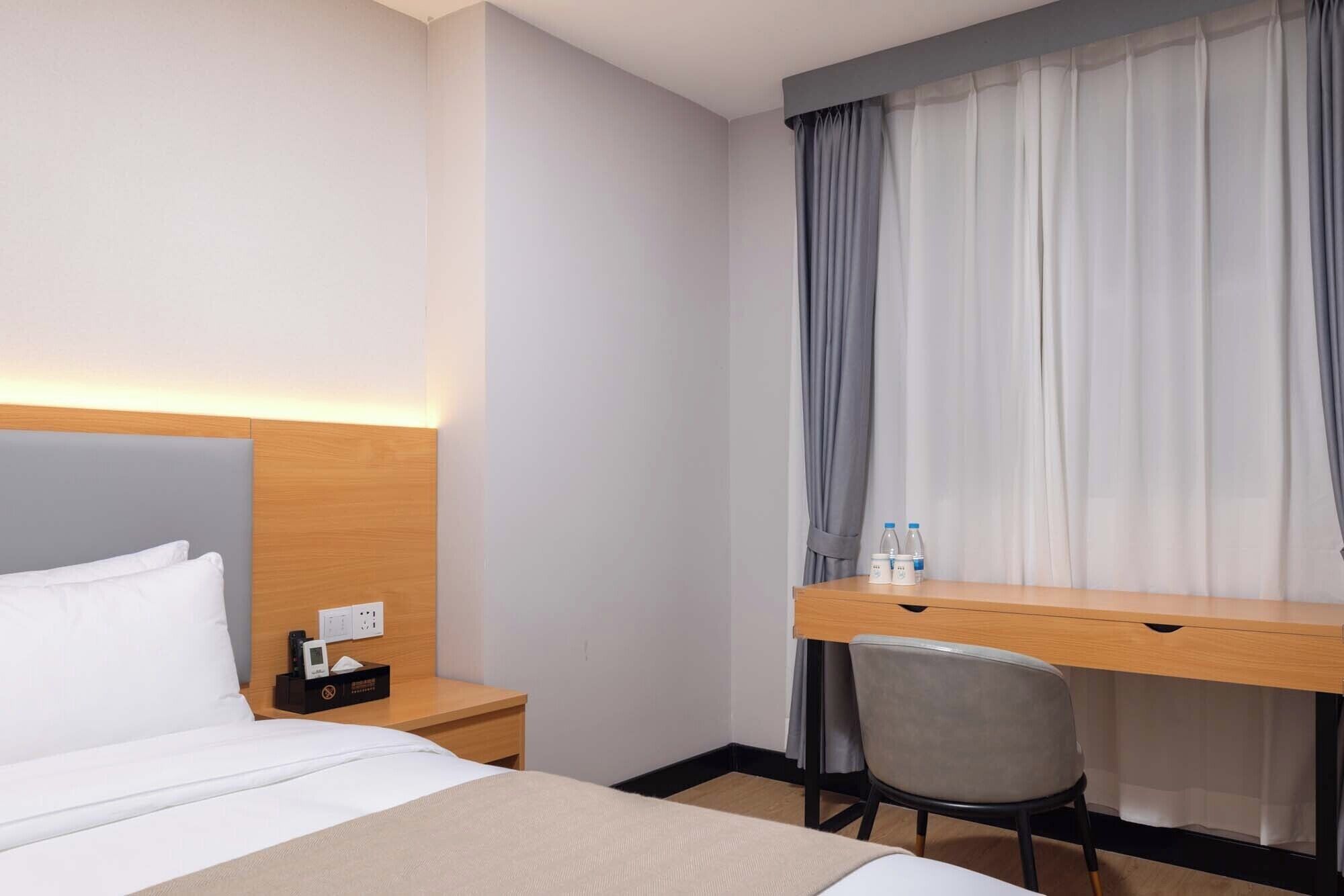 Une chambre d’hôtel avec un lit, un bureau et une chaise. Il y a une fenêtre avec des rideaux, une table de chevet avec un téléphone et une boîte de mouchoirs, et une prise électrique au mur.