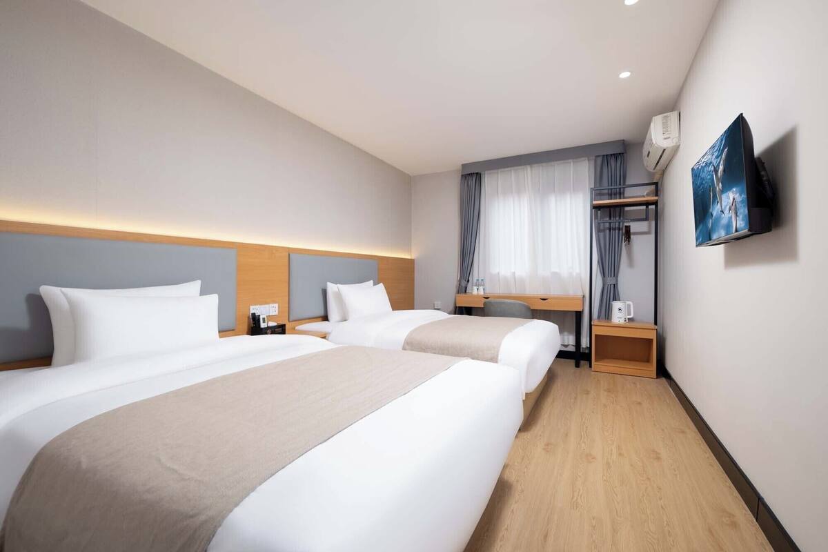 A hotel room with two beds, a desk, a chair, a TV, and an air conditioning unit.