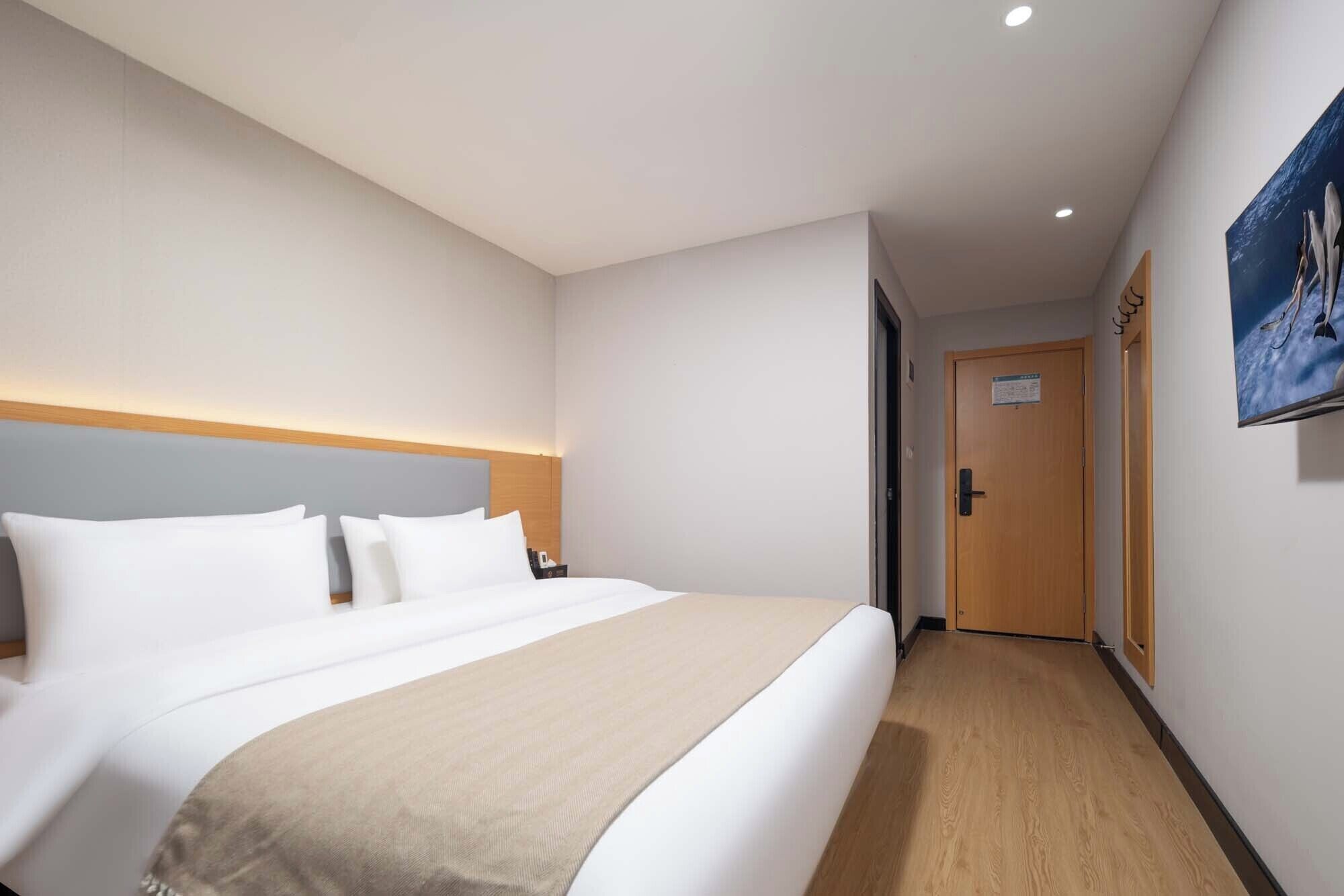 A hotel room with a large bed, a wooden headboard, a beige throw blanket, and a door with a sign.
