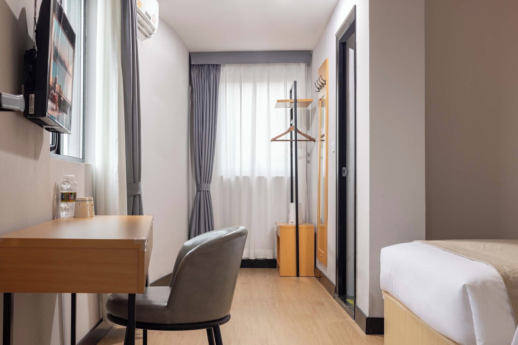 Ett hotellrum med en säng, ett skrivbord, en stol, en klädställning och ett fönster med gardiner.