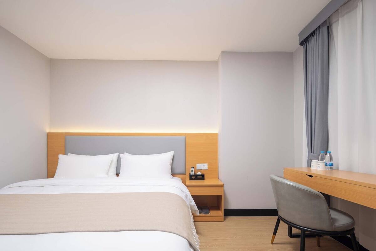 A hotel room with a bed, a desk, a chair, and a nightstand with a phone and a bottle.