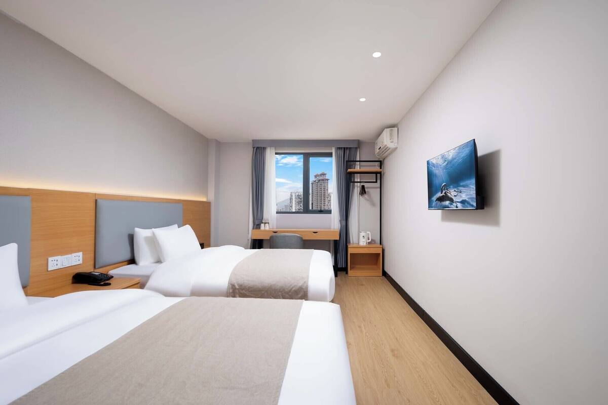A hotel room with two beds, a desk, a chair, a TV mounted on the wall, and a window with a city view.