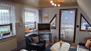 Un salon moderne avec un canapé en cuir noir, une table basse en bois, un téléviseur à écran plat et une vue sur l’extérieur grâce à une porte-fenêtre coulissante.