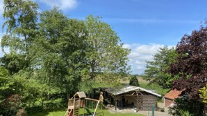 Un espacio al aire libre con césped, un parque infantil de madera, una pequeña estructura cubierta y una casa con techo rojo.