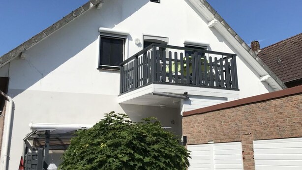 Une maison à deux étages, avec une façade blanche, un balcon doté de rampes noires et une porte de garage.