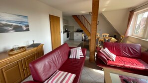 Een ruime woonkamer met een rode leren bank, een houten kast en een eettafel met stoelen. Achtergrond is een zichtbare keuken.