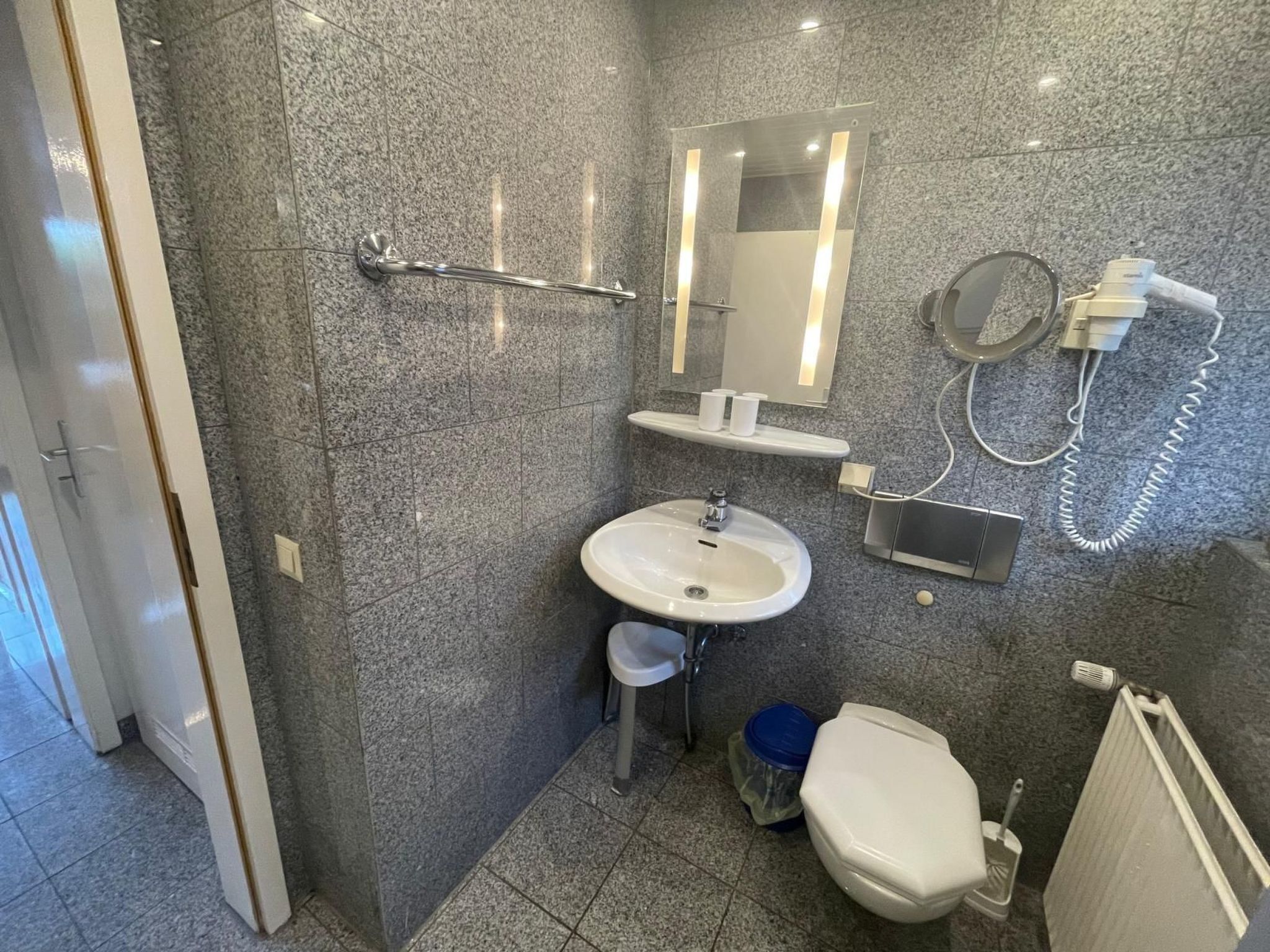 A bathroom with a granite tile wall, a sink, a toilet, a mirror, and a hairdryer.