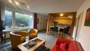 Un espacio habitable compacto con sofá, sillas, una mesa de centro, televisor y una zona de cocina con taburetes de bar.