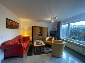 Een woonkamer met een rode bank, een beige fauteuil, een houten kast, een televisie en een raam met uitzicht op groen.