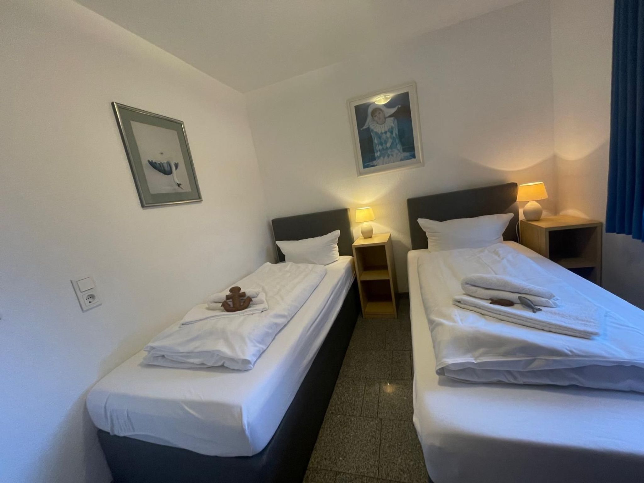 Ein Hotelzimmer mit zwei Einzelbetten, einem kleinen Nachttisch mit Lampe, einem gerahmten Bild an der Wand und einer Steckdose.