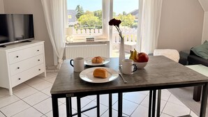 Ein modernes Zimmer mit einem Esstisch, gedeckt mit Speisen, einer Tasse und einer Vase mit Blumen. Ein Fernseher steht auf einem weißen Schrank, und ein Fenster mit Vorhängen ist zu sehen.