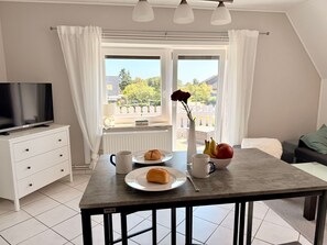 A modern room with a dining table set with a plate of food, a mug, and a vase with flowers. A television is on a white cabinet, and a window with curtains is visible. - Diekbude by Interhome (Friedrichskoog)