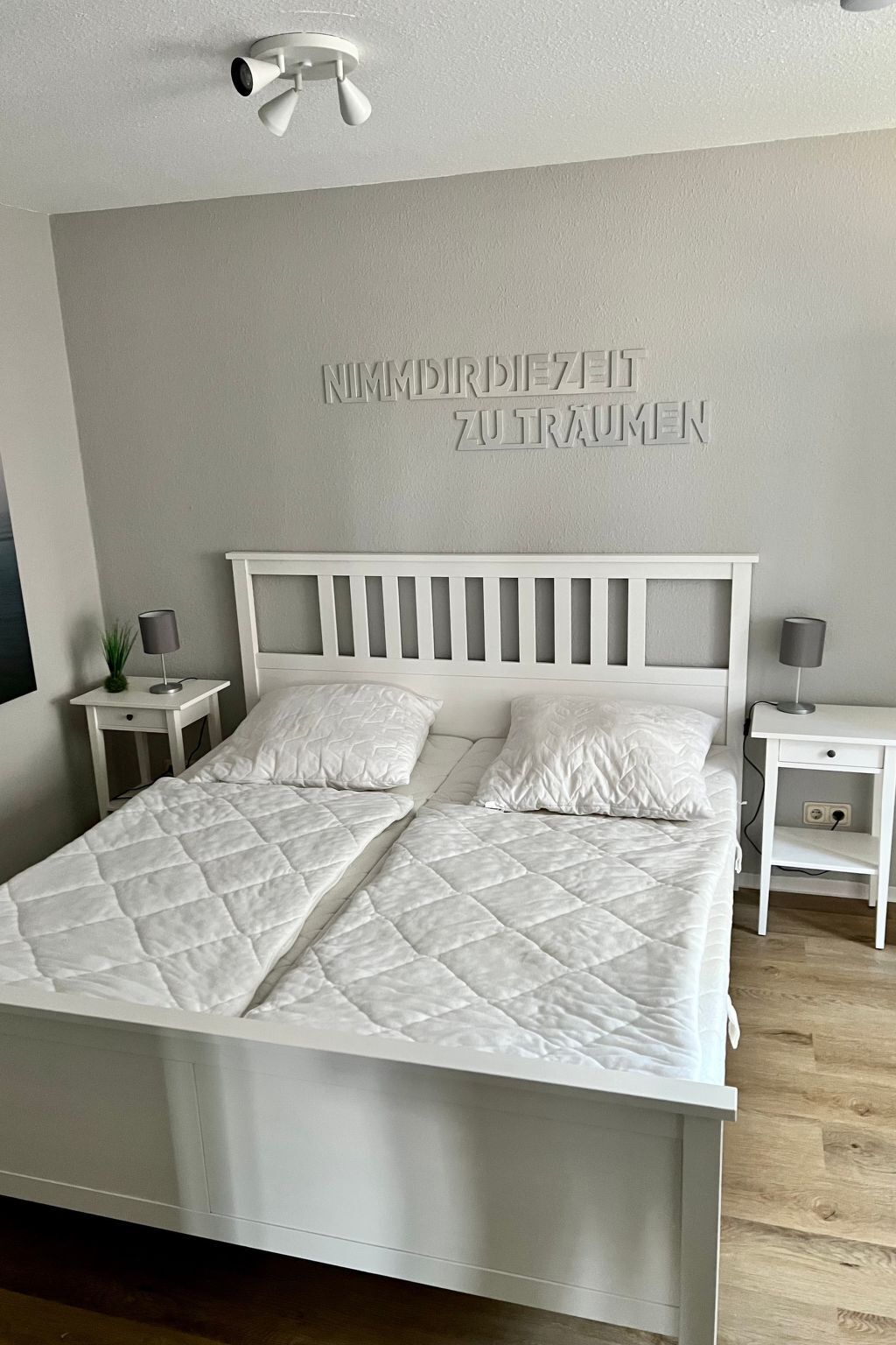 A bedroom with a white bed, two bedside tables, a wall-mounted light fixture, and a wall with the text "NIMMDRIEZEIT ZUTRAUMEN".