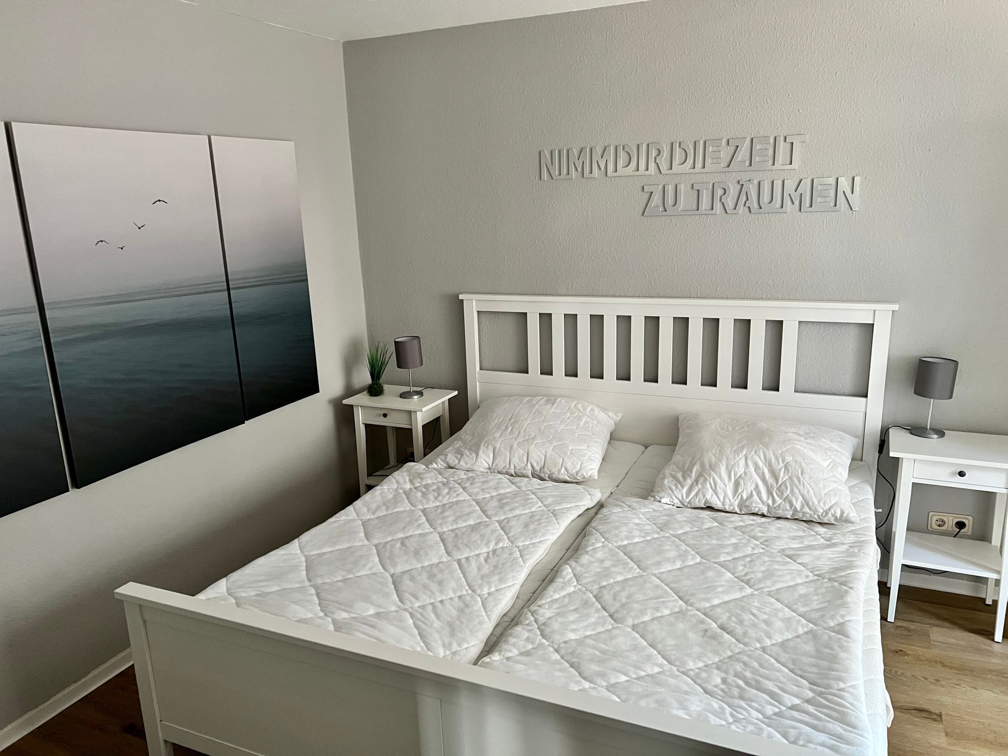 Ein modernes Schlafzimmer mit einem weißen Bett, zwei Nachttischen und einem Wandbild, das Vögel über Wasser zeigt.