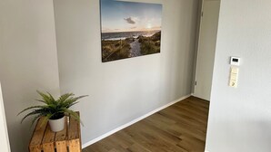 A hallway with a wooden floor, a wall-mounted picture of a beach scene, a potted plant, and a light switch. - Nordseeperle by Interhome (Cuxhaven)