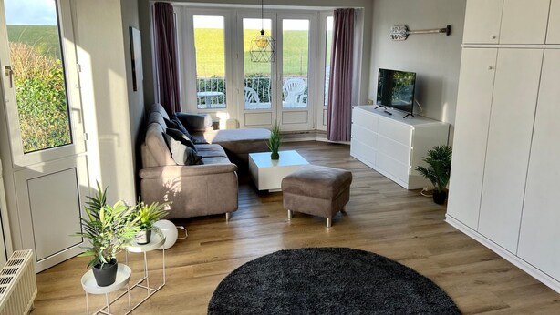A modern living room with a sofa, ottoman, and a TV on a cabinet. There is a balcony with curtains and a view of greenery. - Nordseeperle by Interhome (Cuxhaven)