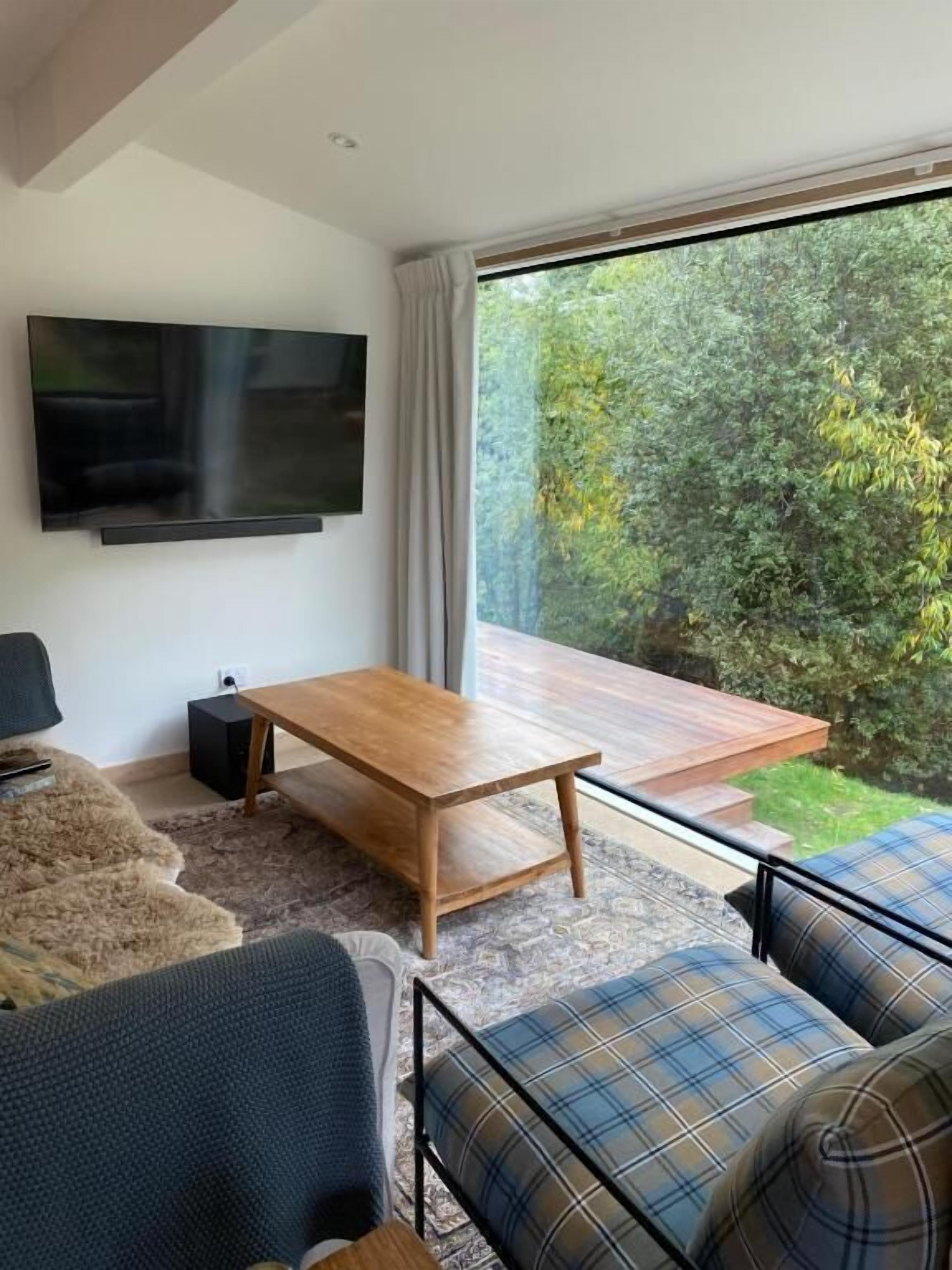 Una sala de estar moderna con un televisor de pantalla plana, una mesa de centro de madera, un sofá y vistas a un jardín a través de amplios ventanales.
