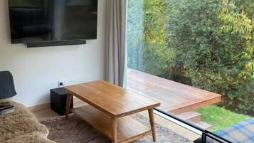 Una sala de estar moderna con un televisor de pantalla plana, una mesa de centro de madera, un sofá y vistas a un jardín a través de amplios ventanales.