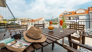 Ein Holztisch mit einem Strohhut, einem Orangensaftglas und einer Zeitschrift auf einem Balkon mit Blick auf eine Stadtlandschaft.