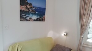 Une pièce avec un canapé vert, un coussin, un tableau représentant un paysage côtier, une fenêtre avec des rideaux et un plancher en bois.
