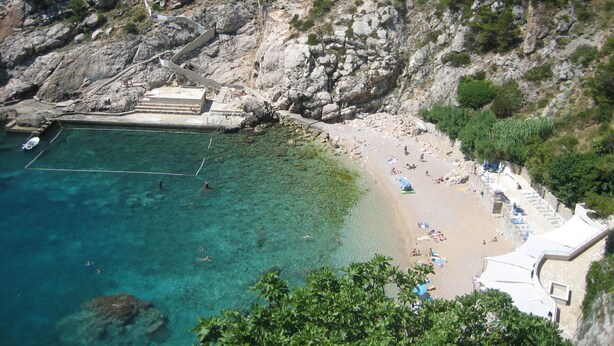 Une plage côtière aux eaux cristallines, bordée de falaises rocheuses, avec quelques bâtiments visibles au loin.