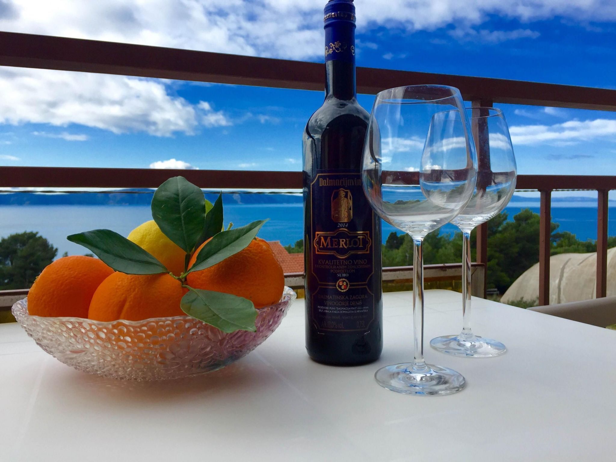 Una ampolla de vi Merlot, dues copes de vi buides i un bol de taronges a un balcó amb una vista panoràmica.