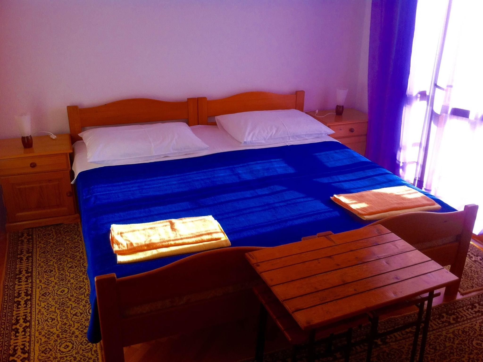 A hotel room with a wooden bed, two pillows, a blue blanket, a wooden table, and a chair.