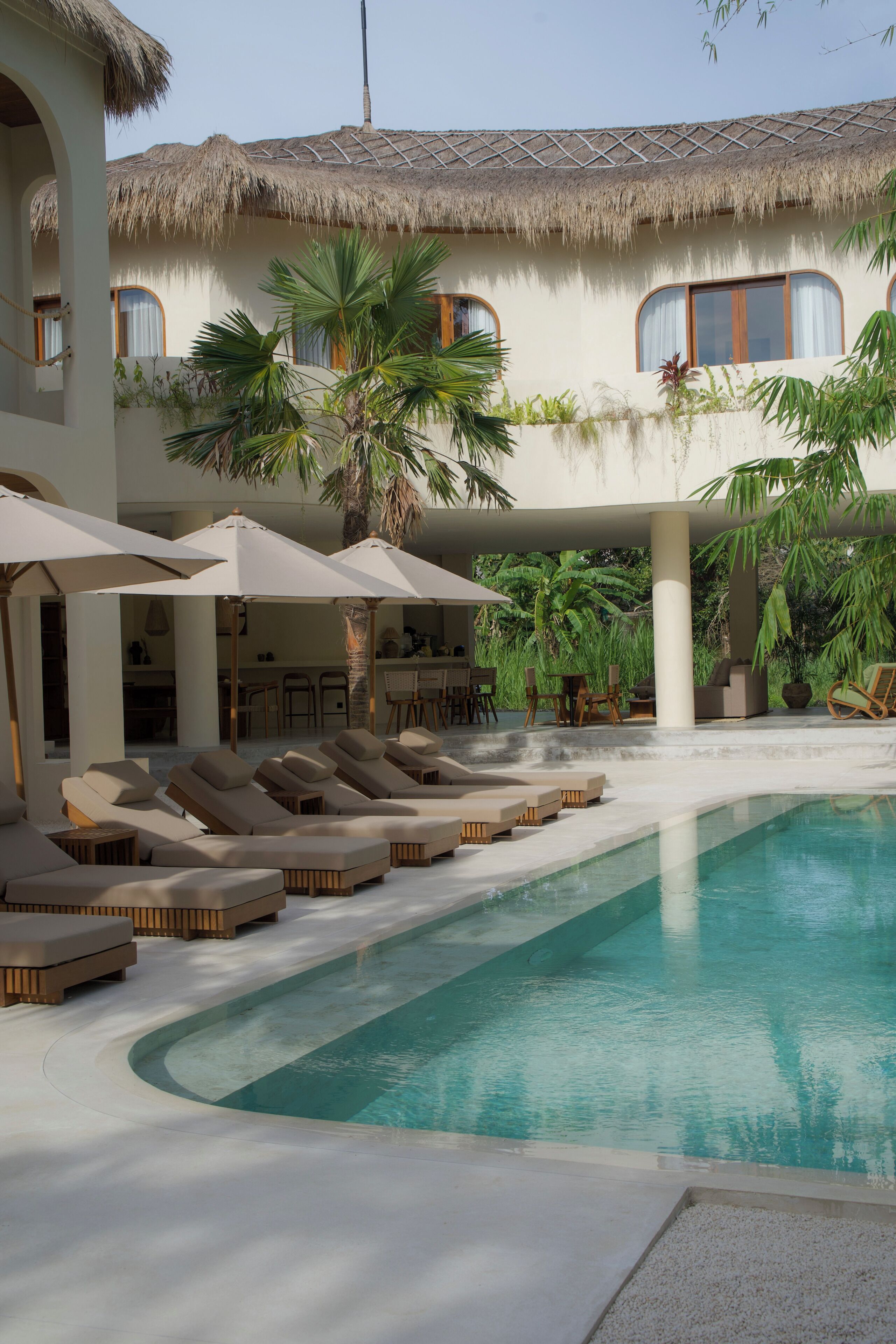 A pool area with lounge chairs and umbrellas, a thatched-roof building, and palm trees.