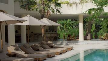 A pool area with lounge chairs and umbrellas, a thatched-roof building, and palm trees.
