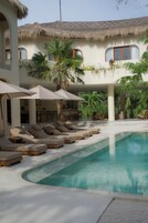 A pool area with lounge chairs and umbrellas, a thatched-roof building, and palm trees.