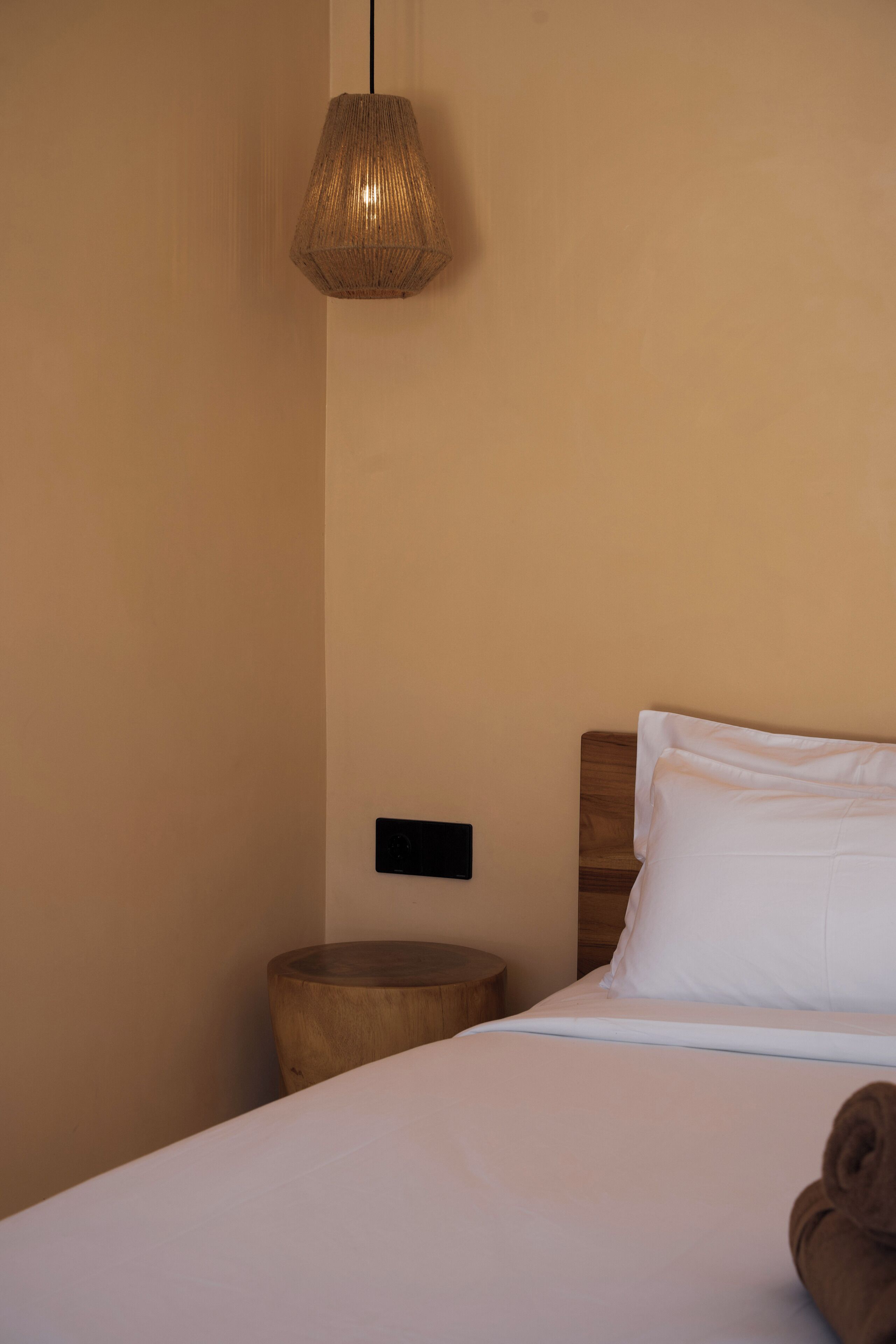 A neatly made bed with white linens, a wooden bedside table, a wall-mounted light fixture, and a small round nightstand.