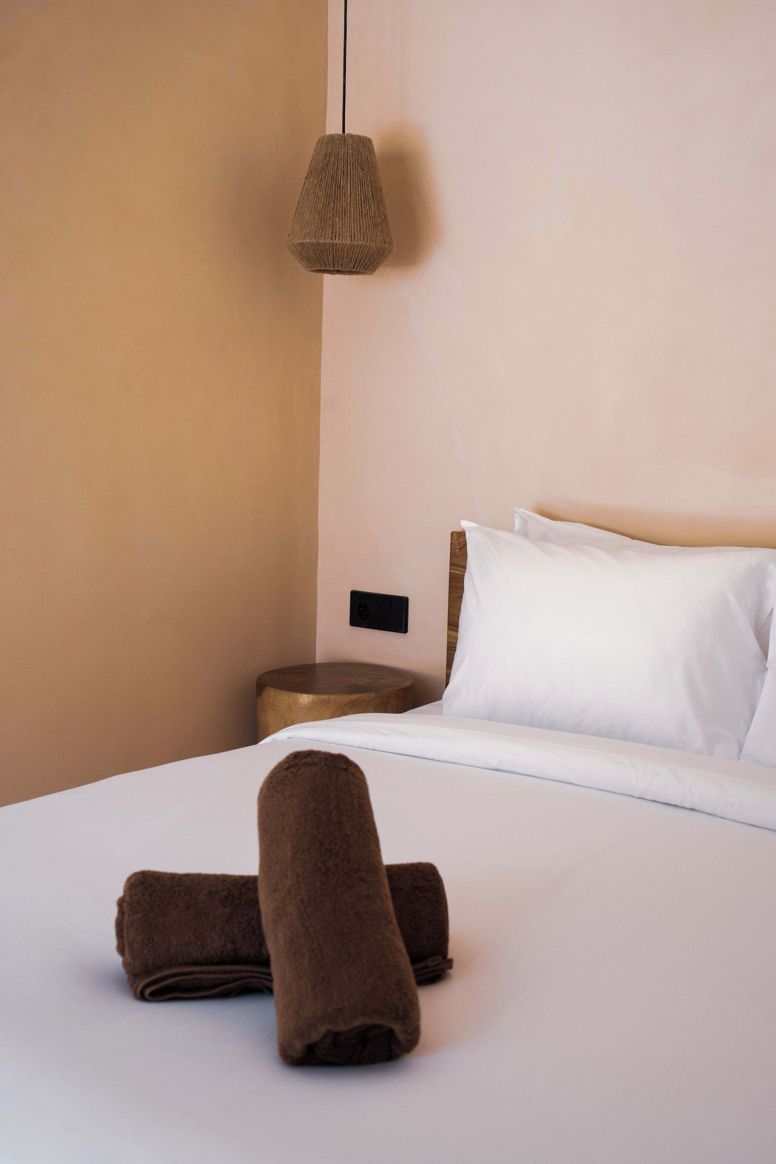 Un lit bien fait, avec du linge de lit blanc et deux serviettes marron, une lampe de chevet et une table de chevet en bois.