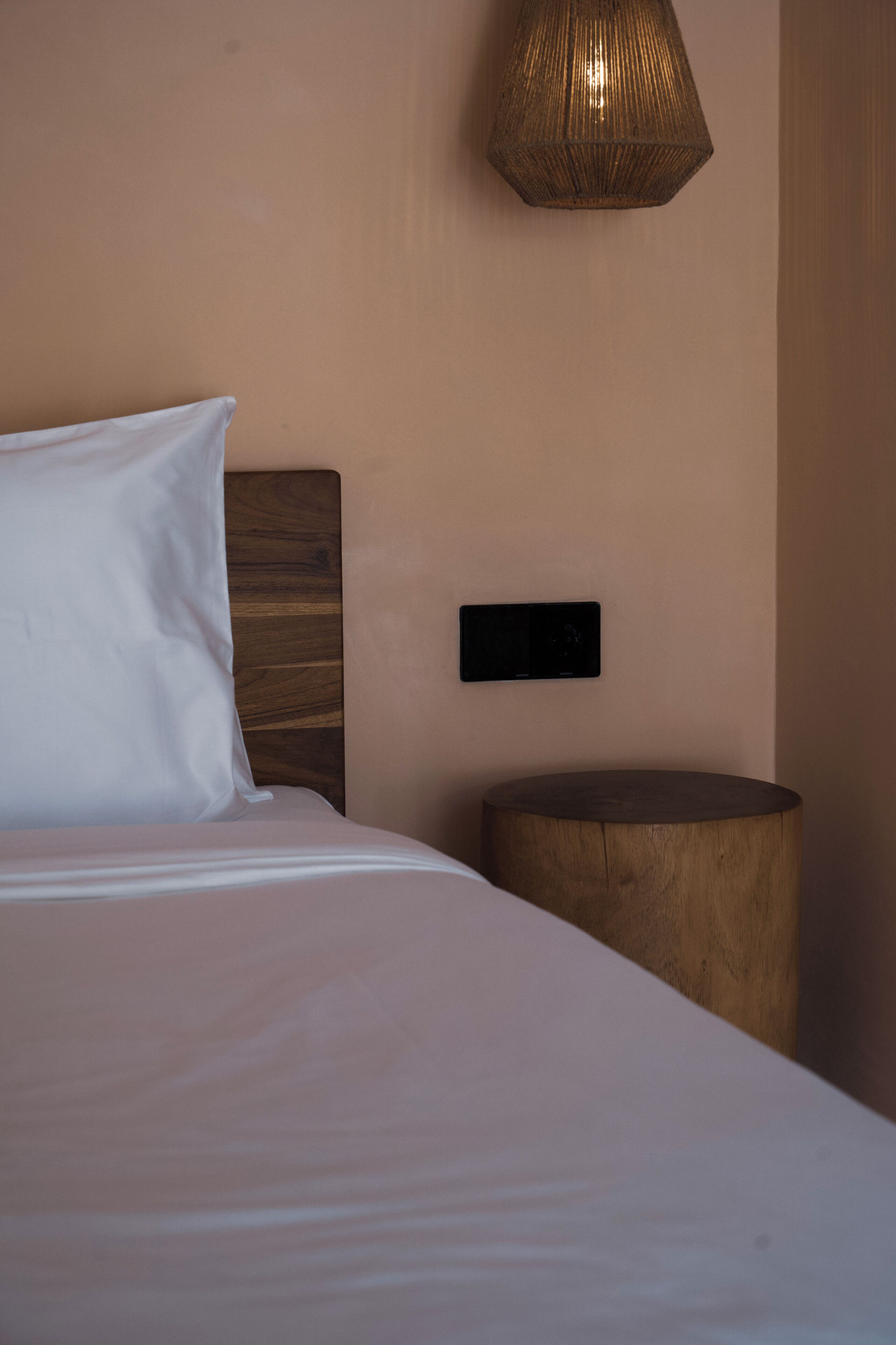 A bed with a pillow, a bedside table, a wall-mounted lamp, and a small round wooden stool.