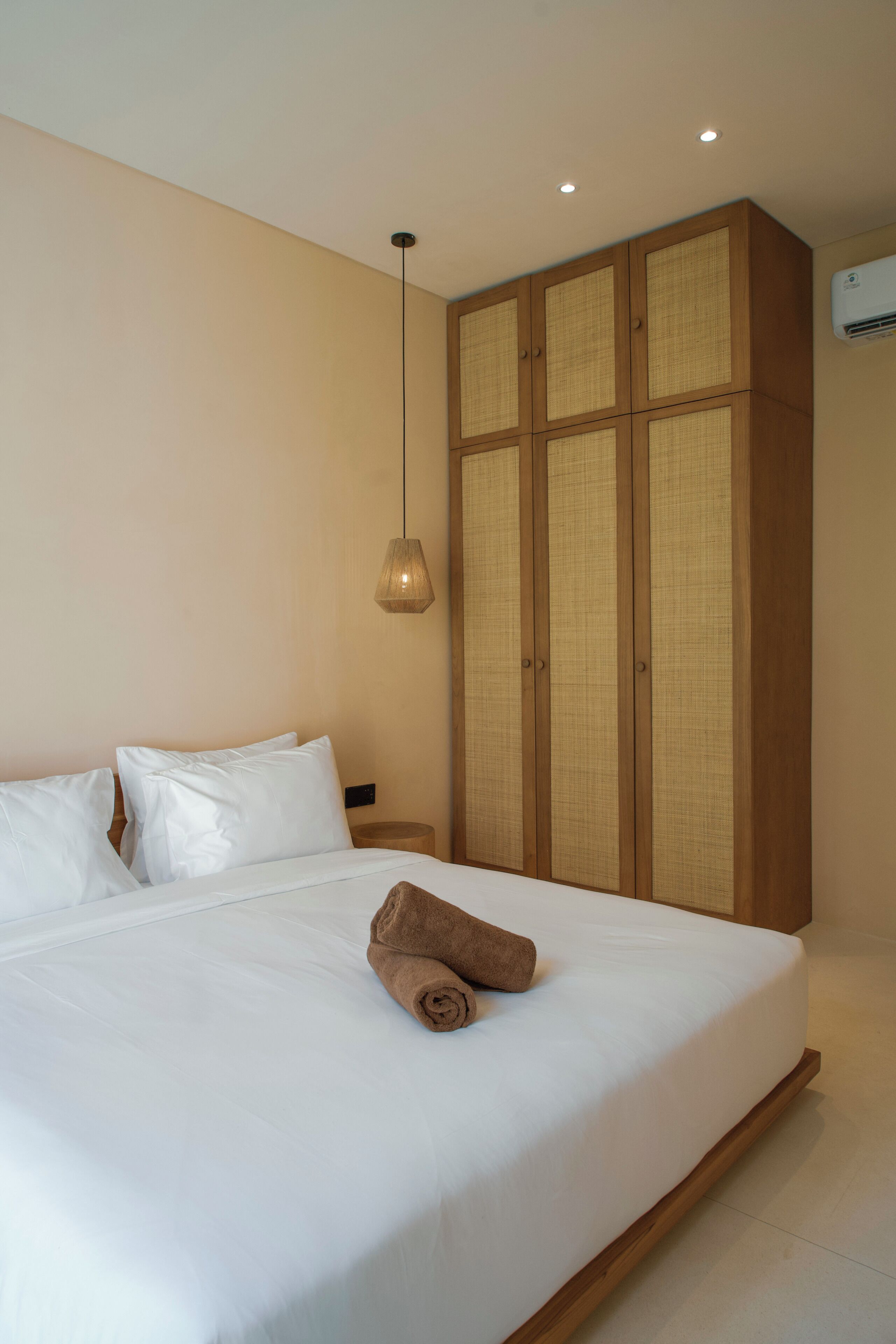 A bedroom with a large bed, a wooden wardrobe, a hanging light fixture, and an air conditioning unit.