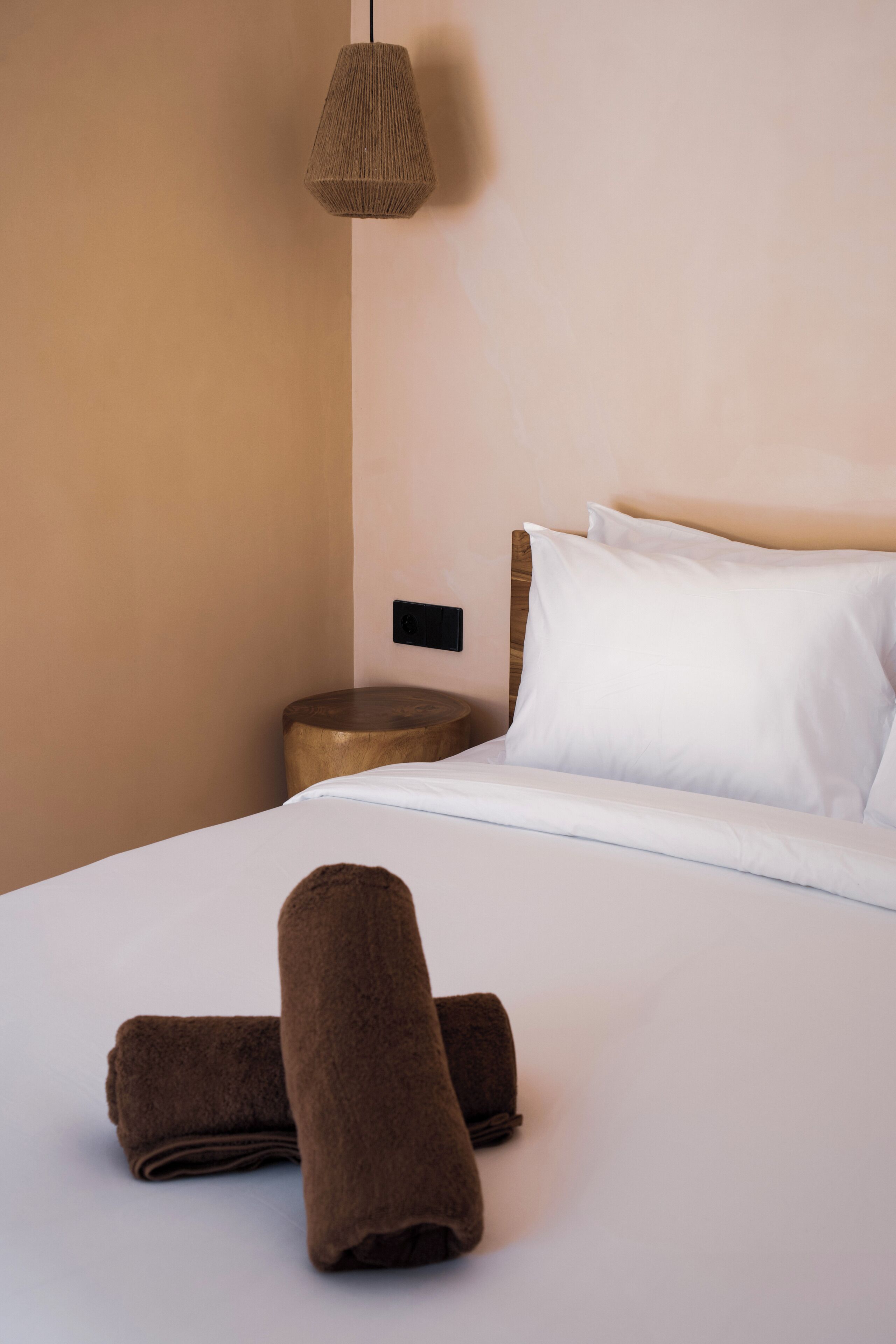 A neatly made bed with white linens, a brown towel, and a wooden nightstand with a lamp.