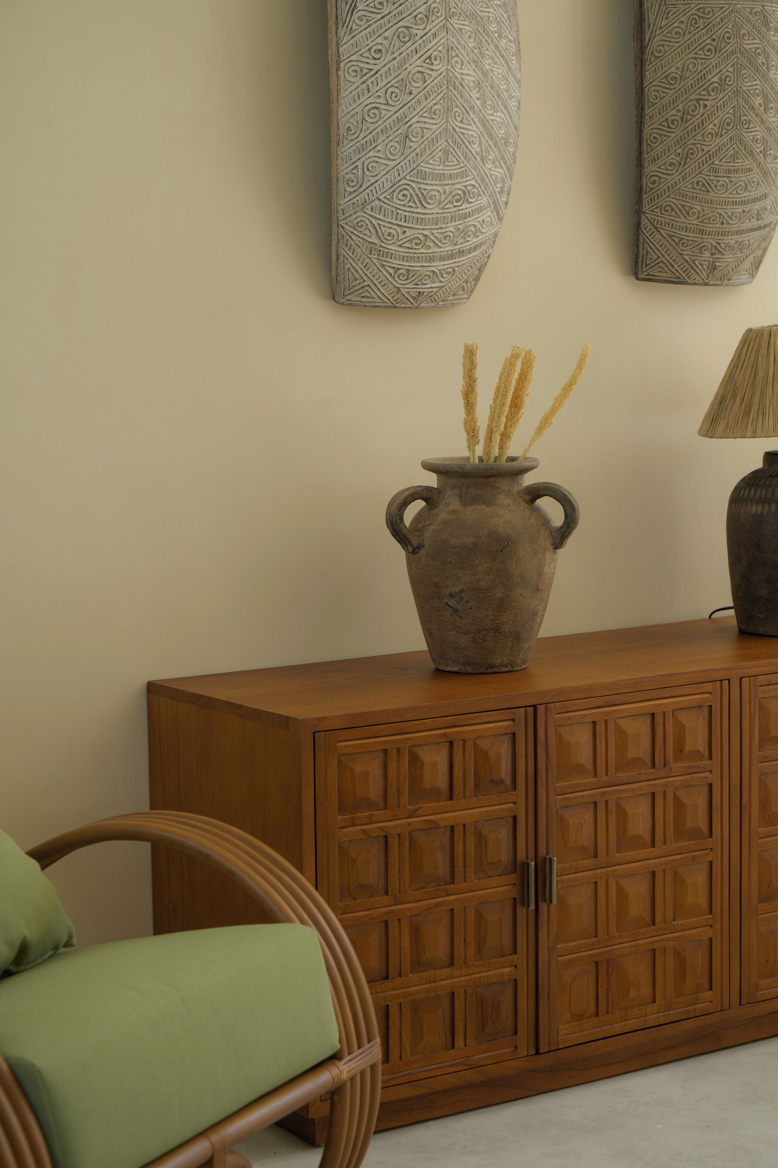 Un mueble de madera con un jarrón de plantas secas, una lámpara y una silla de mimbre.