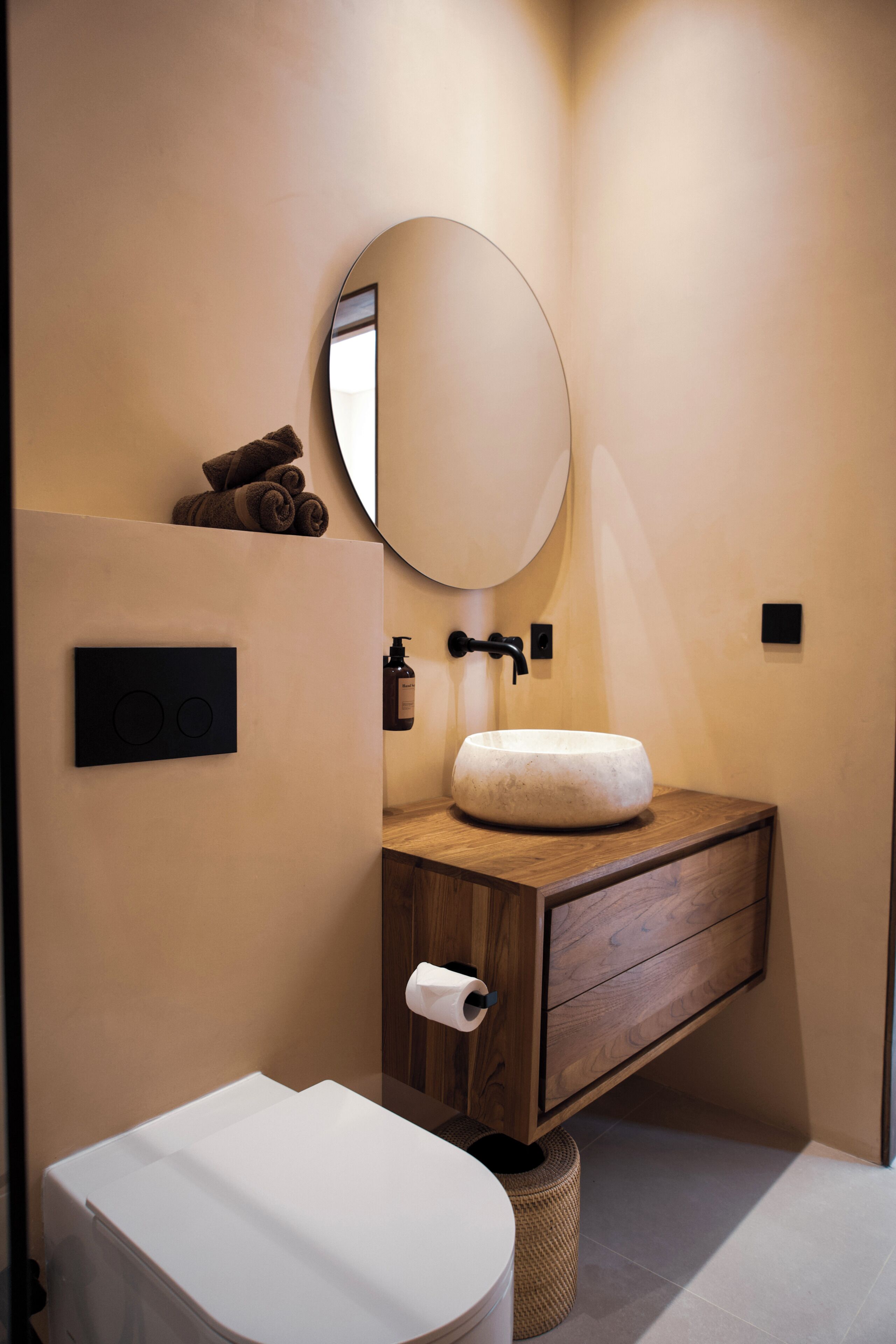 A modern bathroom with a round mirror, wooden vanity, white sink, and a toilet.