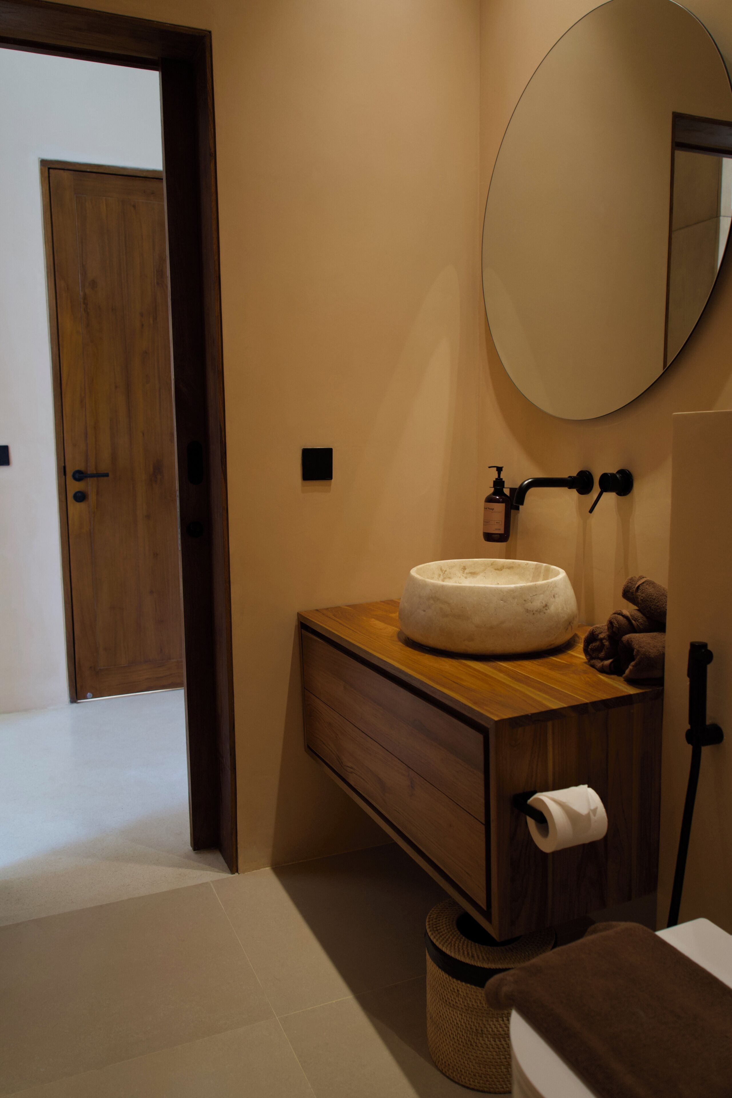 A bathroom with a wooden vanity, a round mirror, a white sink, a towel, and a toilet paper holder.