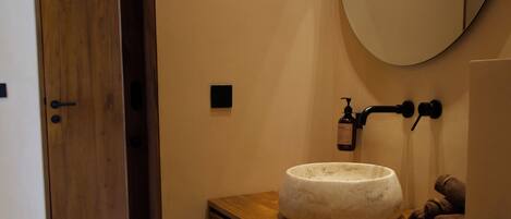 A bathroom with a wooden vanity, a round mirror, a white sink, a towel, and a toilet paper holder.